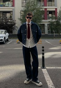 Niebieska dżinsowa kurtka na białej koszuli i czerwonym krawacie, w zestawie z ciemnoniebieskimi dżinsami i czarnymi sneakersami. Sceneria na świeżym powietrzu z budynkami w tle.