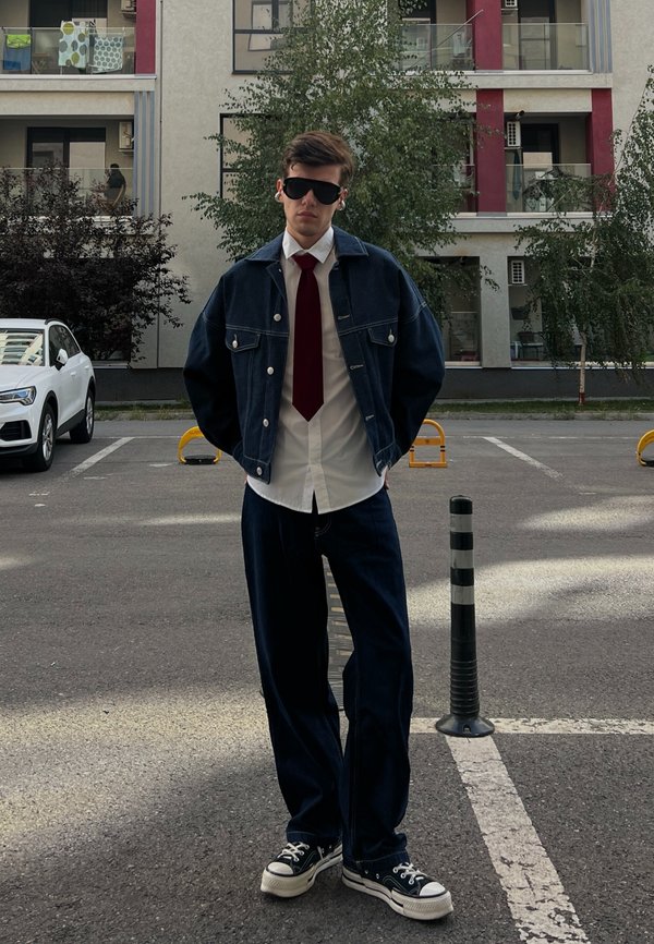 Jachetă din denim albastru peste o cămașă albă și cravată roșie, purtată cu blugi din denim închis și adidași negri. Locație în aer liber cu clădiri în fundal.