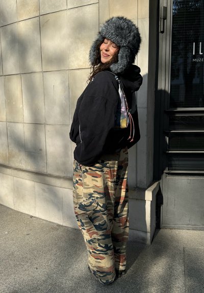 Mujer con sudadera con capucha negra, gorro de piel gris y pantalones de camuflaje, de pie con los ojos cerrados y sonriendo contra una pared con azulejos a la luz del sol.