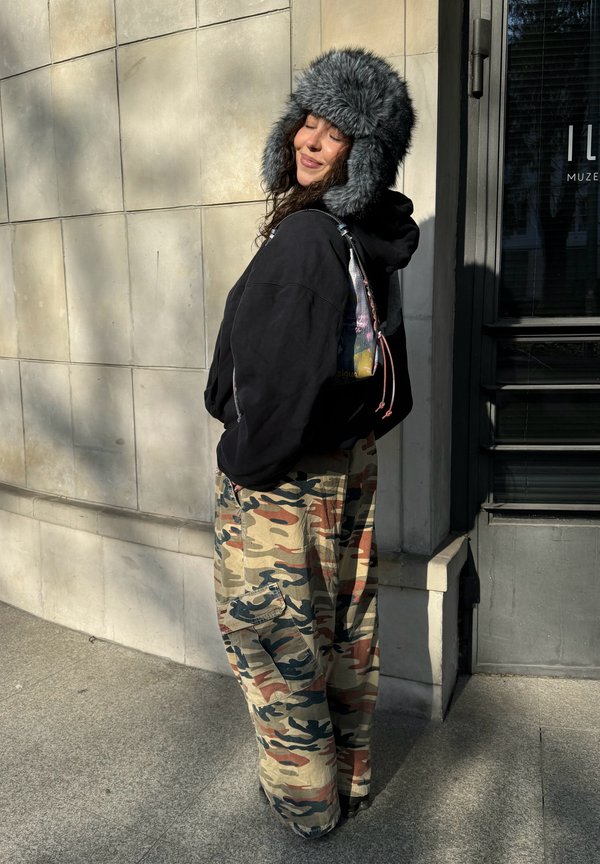 Femme portant un sweat à capuche noir, un chapeau en fourrure gris et un pantalon camouflage, debout les yeux fermés et souriante devant un mur carrelé au soleil.