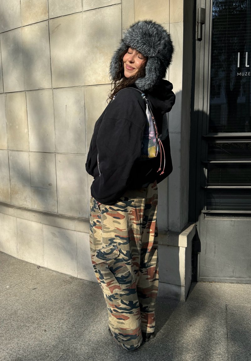 Femme portant un sweat à capuche noir, un chapeau en fourrure gris et un pantalon camouflage, debout les yeux fermés et souriante devant un mur carrelé au soleil.