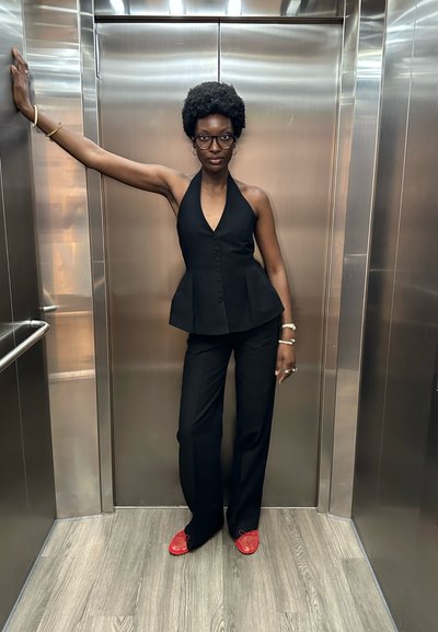 Top halter negro con botones, combinado con pantalones negros y zapatos rojos texturizados. De pie en un ascensor con paredes de acero inoxidable.