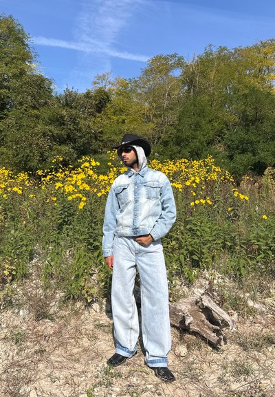 Chaqueta de mezclilla azul claro con dos bolsillos delanteros y capucha gris, combinada con pantalones anchos de mezclilla azul claro. Se lleva un sombrero de vaquero negro y gafas de sol.