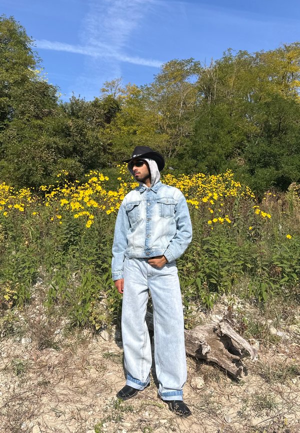 Lichtblauwe denim jack met twee voorzakken en grijze capuchon, gecombineerd met lichtblauwe wijde jeans. Zwarte cowboyhoed en zonnebril gedragen.