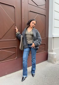 Oversized mørkegrå ulljakke over en grønn omslagsgenser, kombinert med vide blå jeans og svarte spisse hæler, foran en brun dør.