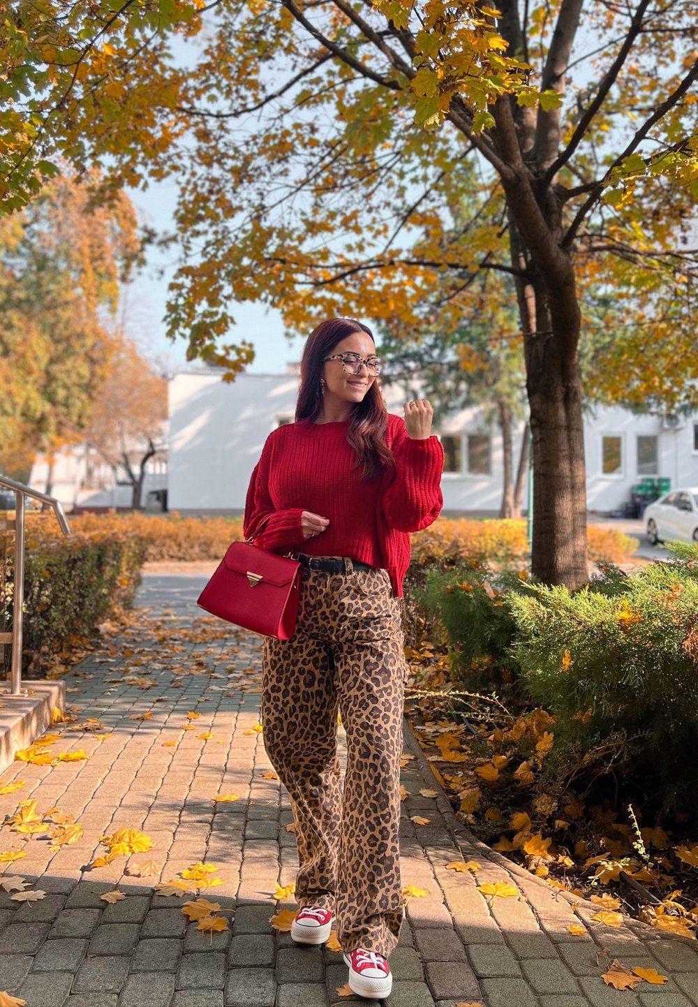 Pulover tricotat roșu, pantaloni cu imprimeu leopard și adidași albi cu accente roșii. Ținând o geantă roșie; frunze de toamnă în fundal.