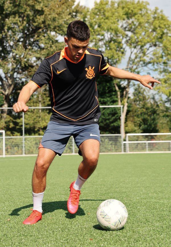 Svart sportsdrakt med oransje detaljer, grå shorts og røde fotballsko. Spiller dribler en mønstret fotball på grønt kunstgress.