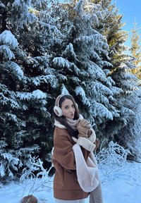 Jachetă din suede căprui cu accente de blană albă, eșarfă bej, mănuși căprui și căști albe de urechi, așezată pe fundalul pomilor verzi acoperiți de zăpadă.