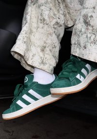 Grønne sneakers i semsket skinn med hvite striper, brukt med hvite sokker og lyse mønstrete bukser, føttene hviler på en mørk overflate.