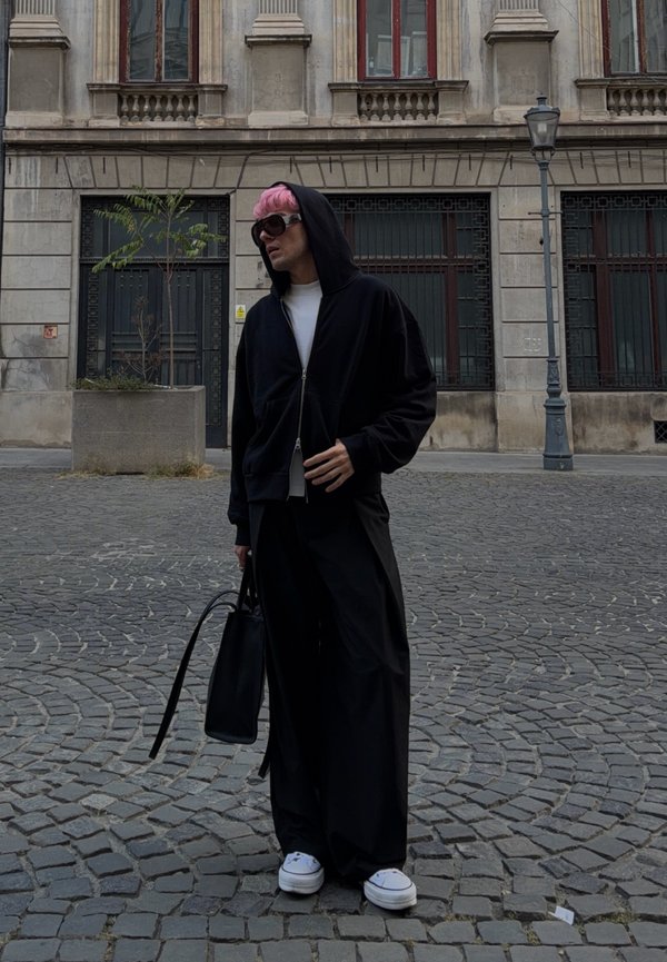 Persona con pelo rosa y gafas de sol, vistiendo una sudadera negra con capucha y pantalones anchos, sosteniendo una bolsa negra, de pie en una calle empedrada.