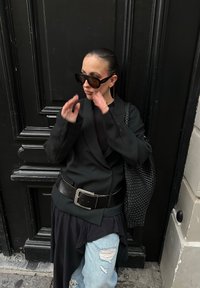 Femme portant des lunettes de soleil surdimensionnées, un blazer noir avec une large ceinture, un jean clair déchiré, et tenant un sac noir tissé, debout devant une porte noire.