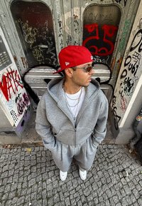 Szürke zipzáros kapucnis pulóver és ahhoz illő nadrág, elülső zsebekkel. Piros sapkával és fehér tornacipővel viselve. Városi háttér graffiti-val.