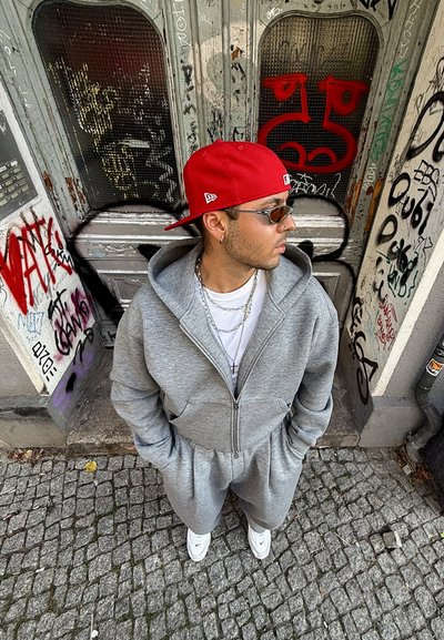 Sudadera gris con cremallera y pantalones a juego, que cuentan con una capucha y bolsillos frontales. Combinada con una gorra roja y zapatillas blancas. Fondo urbano con graffiti.