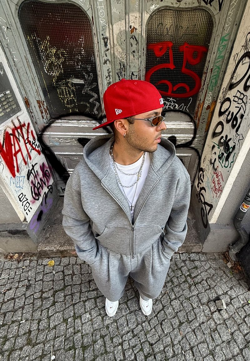 Grey zip-up sweatshirt and matching trousers, featuring a hood and front pockets. Worn with a red cap and white trainers. Urban backdrop with graffiti.