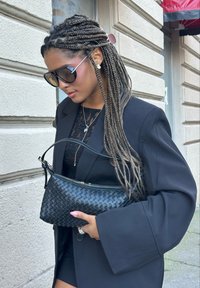 Czarna pleciona skórzana torba o zaokrąglonym kształcie, z zamkiem błyskawicznym i pojedynczym uchwytem. Noszona z czarną, oversize'ową marynarką i okularami przeciwsłonecznymi.