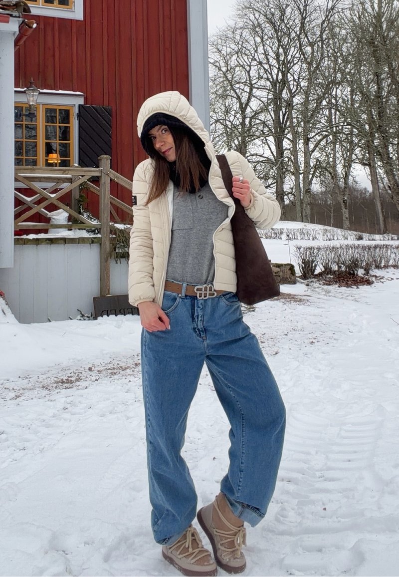 Donna con giacca invernale e jeans in piedi sulla neve vicino a una casa di legno rossa, che tiene in mano una borsa marrone con alberi senza foglie sullo sfondo.
