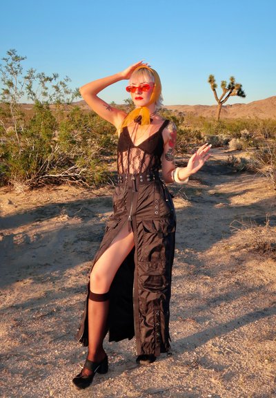 Persona con cabello rubio, pañuelo naranja en la cabeza, gafas de sol rojas, blusa negra de encaje y falda larga negra, de pie en un paisaje desértico con arbustos y un árbol.