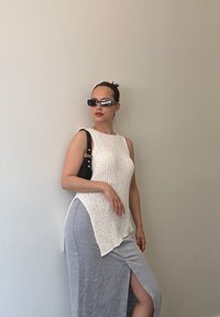 Bela pletena brezrokavna majica z asimetričnim robom, v paru z svetlo sivo maksi krilo z bočnim razporkom. Dodatki vključujejo črno torbico in sončna očala.