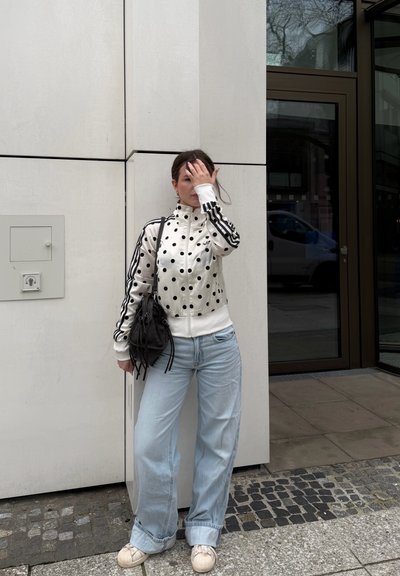 Mujer con chaqueta blanca de lunares, vaqueros anchos azul claro, zapatillas blancas, sosteniendo bolso negro, de pie contra una pared beige afuera de un edificio.