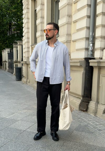 Camisa de botones a rayas, camiseta blanca debajo, pantalones negros, zapatos negros y una bolsa de mano beige; de pie en una acera de la ciudad cerca de una arquitectura histórica.