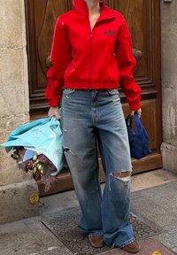 Personne portant une veste rouge zippée et un jean large déchiré, tenant un bouquet enveloppé dans du papier bleu dans une main et un sac bleu dans l'autre.