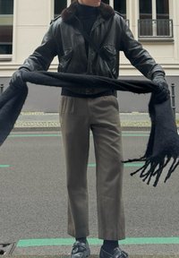 Veste bombardier en cuir noir avec col en fourrure, écharpe noire, pantalon gris et mocassins noirs. Le matériau est lisse avec une coupe structurée.
