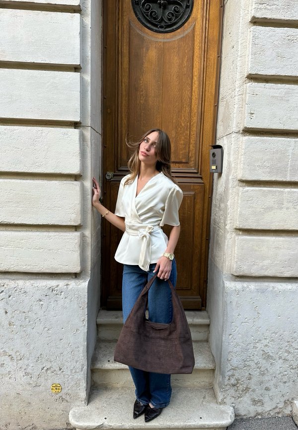 Mujer con blusa blanca y vaqueros sosteniendo una gran bolsa marrón, de pie sobre escalones de piedra frente a una puerta alta de madera con interfono en un edificio de piedra.