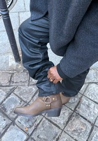 Bottine en cuir marron à bout rond, avec une bride décorative et des accents dorés, portée avec un pantalon noir, sur une surface en pierre texturée.