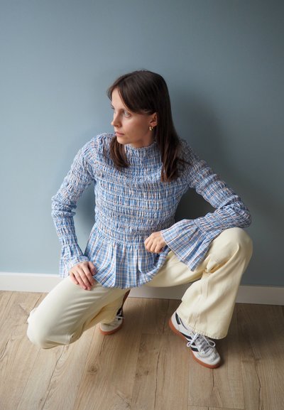 Camisa a cuadros azul y blanca con diseño fruncido, de manga larga y puños acampanados. Combinada con pantalones crema y zapatillas deportivas, sobre un suelo de madera.