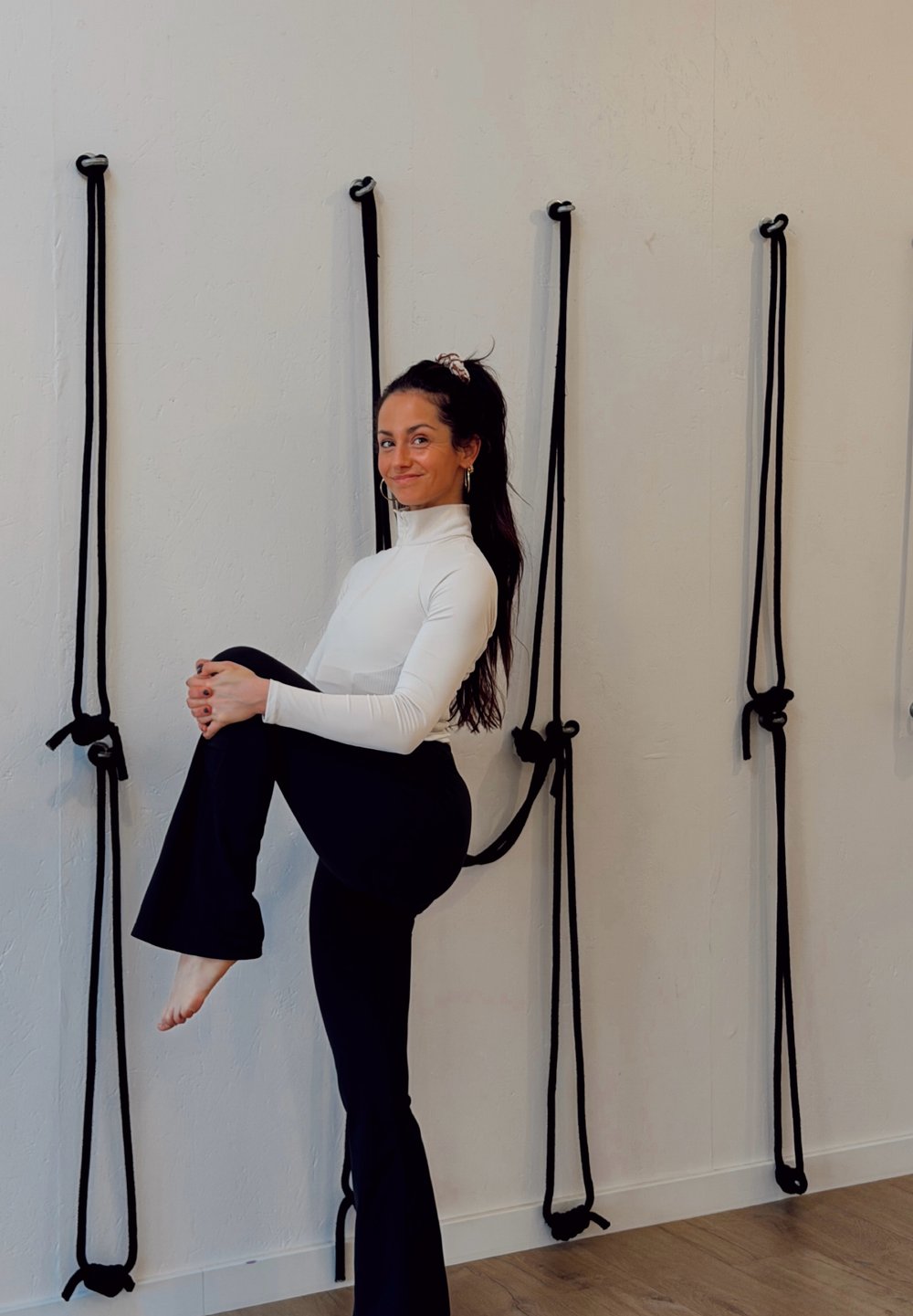 Femme portant un haut à manches longues blanc et un pantalon évasé noir, levant un genou. Des cordes noires sont attachées à un mur clair derrière elle.