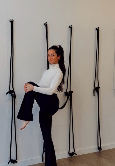 Mujer con una blusa blanca de manga larga y pantalones acampanados negros, levantando una rodilla. Cuerdas negras están sujetas a una pared de color claro detrás de ella.