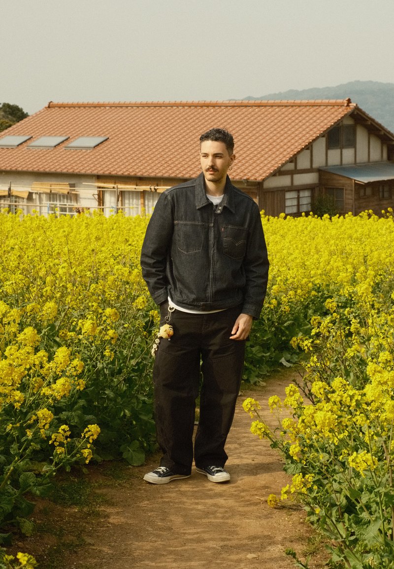 Homme avec veste sombre et pantalons noirs debout sur un chemin en terre entouré de plantes à fleurs jaunes, avec une maison au toit en tuiles en arrière-plan.