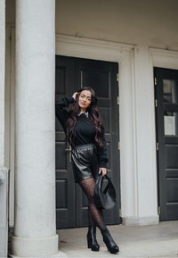 Pull noir, shorts en cuir et collants à motifs. Bottines noires et sac à main. Debout près d'une colonne avec des portes sombres en arrière-plan.