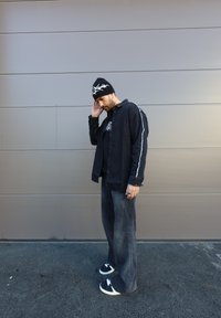Cămășă din denim negru cu cusături albe la mâneci, suprapusă peste un tricou grafic negru. Jeans negri largi și adidași negri cu accente albe.