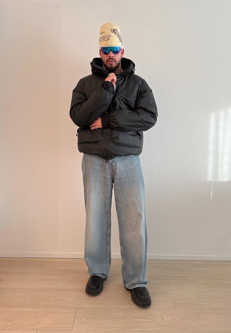 Homme portant un bonnet beige, des lunettes de soleil réfléchissantes, une doudoune noire, un jean bleu clair et des chaussures noires, debout avec la main près de la bouche à l'intérieur.
