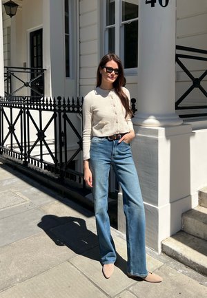Cardigan de punto acanalado en color beige, con botones, combinado con jeans de pierna ancha en azul claro. Bailarinas de punta en color beige. Gafas de sol negras. Entorno urbano.