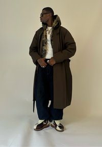 Mann in einem dunkelbraunen langen Mantel, Tarnkapuzenpullover, weißem Hemd, lockeren dunklen Jeans, Rindsleder-Schuhen, Sonnenbrille und mehrlagigen silbernen Halsketten, der seinen Gürtel anpasst.