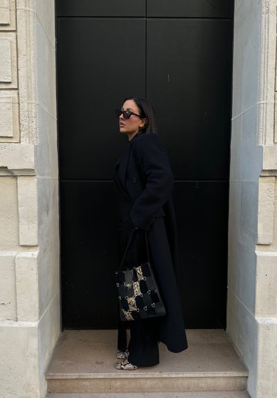 Abrigo largo negro, bolso estampado en negro y beige con acentos metálicos, atuendo negro texturizado y zapatos de piel de serpiente en escaleras de piedra.