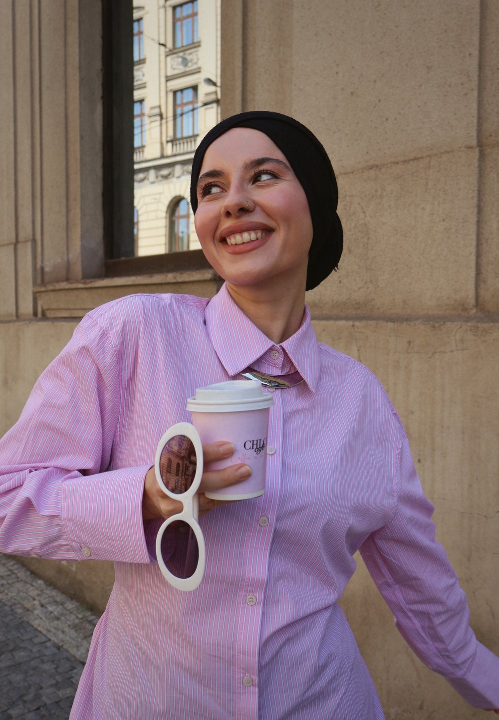 Rosa og hvit stripete skjorte, svart hijab, store hvite solbriller, holder en kaffekopp med et rosa beskyttelseslag, står mot en steinmur.