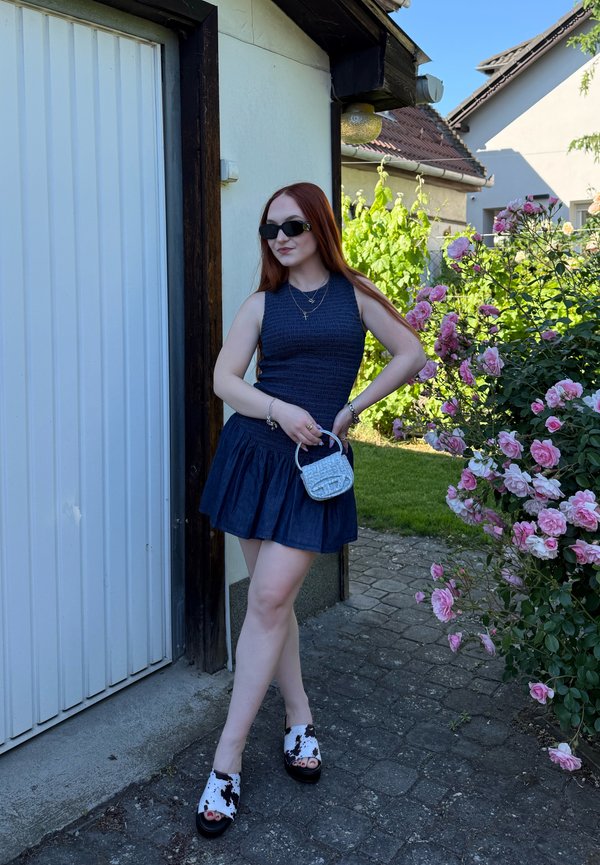 Jeune femme en robe bleu foncé sans manches et lunettes de soleil se tient près de la porte du garage à côté d'un buisson de rosiers roses en fleurs dans un jardin ensoleillé.