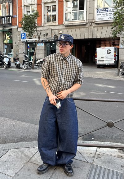 Camisa de cuadros de manga corta, pantalones de mezclilla azules oversized, zapatos oscuros, sosteniendo una lata de bebida, de pie junto a una barandilla de metal en la calle.