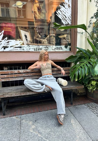 Una joven está sentada en un banco de madera, vistiendo un top corto de cuadros y pantalones de chándal grises holgados, con el cabello ondulado, adornada con anillos y un bolso pequeño.
