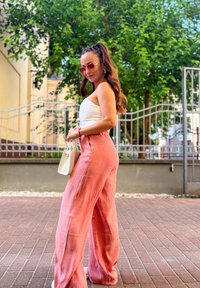 Light pink linen wide-leg trousers with a textured finish, teamed with a white fitted top. Carrying a small white handbag. Green background.