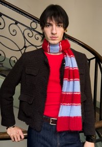 Jeune personne aux cheveux foncés portant une chemise rouge, une veste noire texturée, une écharpe rayée rouge et bleu clair, debout près d'une balustrade noire ornée.