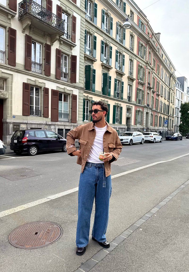 Homem com óculos de sol, casaco castanho, camisa branca e jeans largos azuis, segurando uma bebida, em pé numa rua da cidade com carros estacionados e edifícios.