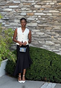 Κρέμ αμάνικο πουκάμισο με χρυσά κουμπιά, μαύρη φαρδιά φούστα, λευκά ψηλοτάκουνα παπούτσια και μία μικρή μαύρη τσάντα με χρυσές λεπτομέρειες, μπροστά από έναν πέτρινο τοίχο.