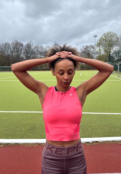 Mujer con una camiseta sin mangas rosa y leggings morados está de pie en un campo deportivo con las manos entrelazadas en la cabeza, los ojos cerrados, preparándose o descansando.
