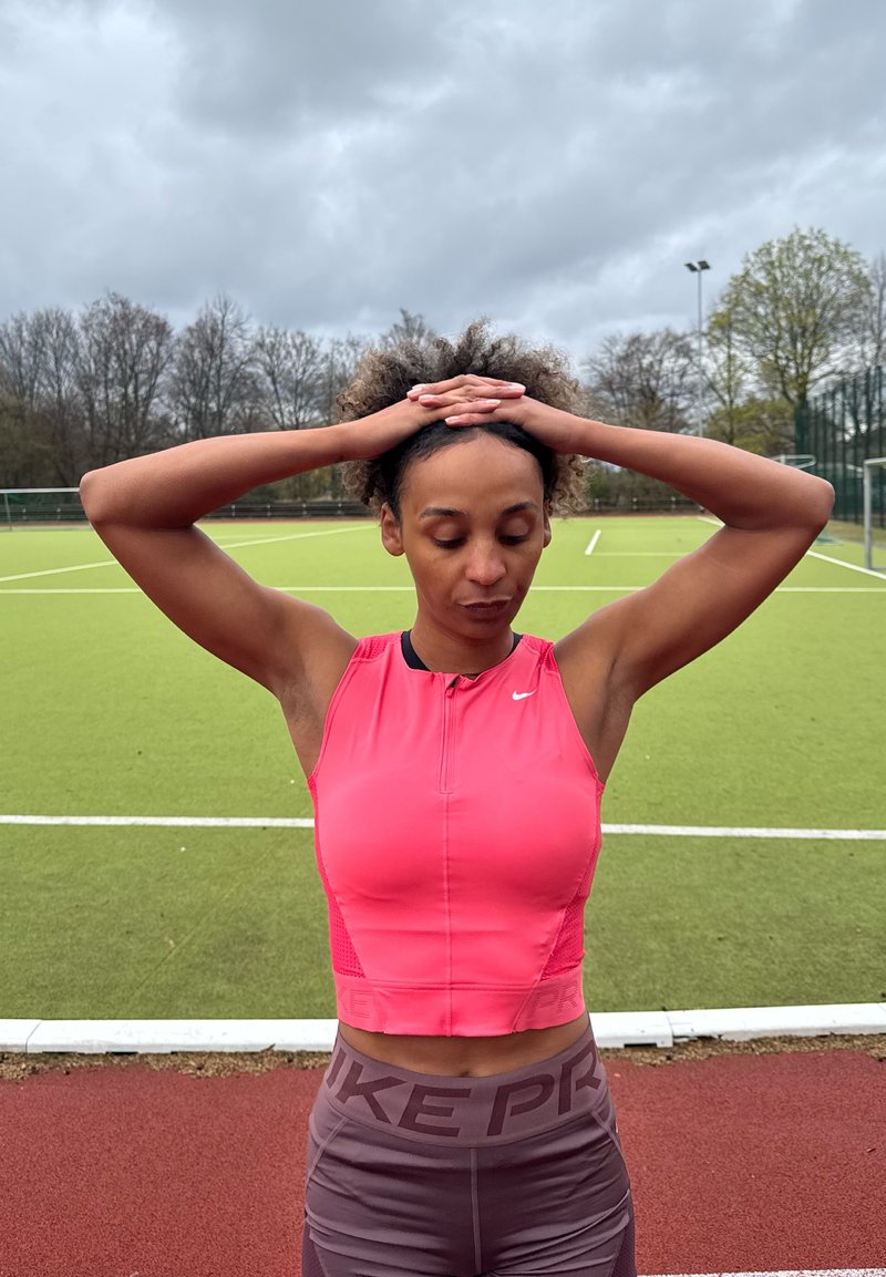 Femme en haut sans manches rose et leggings violets se tenant sur un terrain de sport avec les mains jointes sur la tête, yeux fermés, en train de se préparer ou de se reposer.