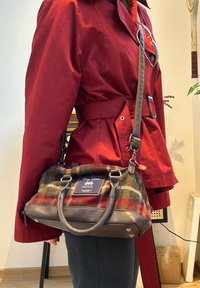 Personne portant une veste rouge ceinturée et un sac à main à carreaux marron et rouge avec des lanières en cuir et une étiquette de la marque MOON en bandoulière.