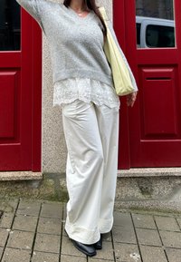 Άτομο που φοράει γκρι πουλόβερ πάνω από λευκό μπλουζάκι με δαντέλα, χαλαρά λευκά παντελόνια, μαύρα παπούτσια και κρατάει μια ανοιχτοκίτρινη τσάντα ώμου, στέκεται δίπλα σε κόκκινες πόρτες.